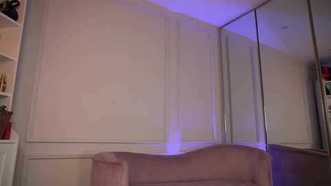 Media: Video of a modern, minimalist bedroom with beige walls, a white wardrobe mirror, and a pink upholstered chair. A subtle purple light highlights the scene, creating a serene ambiance.