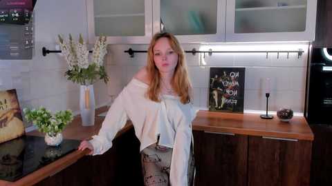 Media: Video of a blonde Caucasian woman in a white off-shoulder shirt, leaning on a modern kitchen counter with wooden cabinets, flowers, and a black decorative sign.