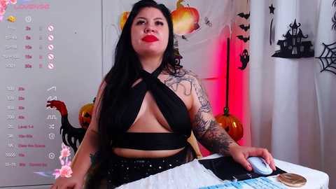 Media: Video of a curvy, tattooed woman with long black hair and red lipstick, wearing a black halter top and high-waisted pants, sitting at a desk with a calendar, pumpkin decor, and a computer.