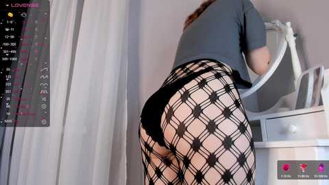 Media: Video of a woman with fair skin wearing black fishnet stockings and a grey top, bending over a white mirror, partially visible.