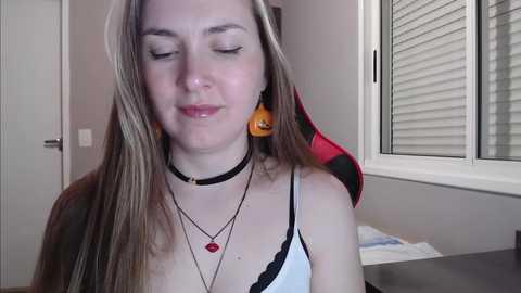 Media: A video of a young woman with long, straight, light brown hair, wearing a black choker and multiple necklaces, including a red heart pendant, in a dimly lit room with a window and red gaming chair in the background.