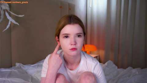 Media: Video of a young woman with fair skin, light brown hair, and brown eyes, sitting on a bed in a dimly lit room. She wears a white shirt and appears contemplative.