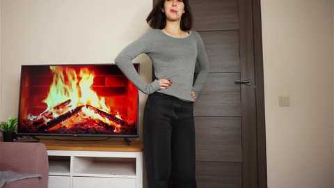 Media: Video of a confident woman with straight brown hair, wearing a gray sweater and black pants, standing in front of a lit fireplace TV on a white cabinet, with a closed wooden door in a modern living room.