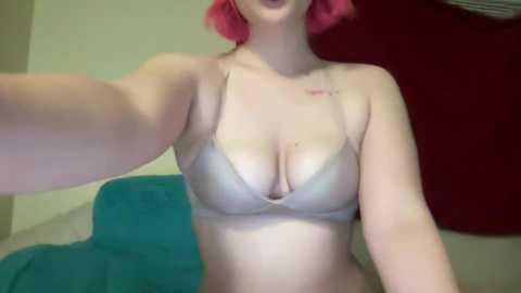 Media: Video of a fair-skinned woman with pink hair, wearing a beige bra, and a tattoo on her left collarbone, sitting on a blue couch in a room with a red wall.