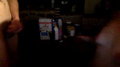 Media: A dimly lit video of two nude, erect penises and a cluttered table with various food items and toiletries in the background.