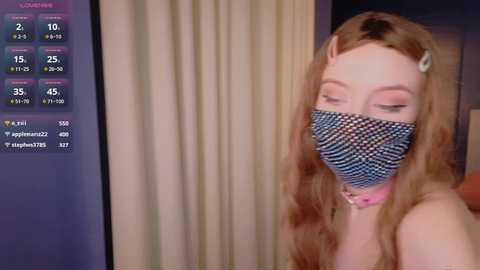 Media: Video of a young woman with long, wavy blonde hair, wearing a blue-and-white checkered face mask and pink collar, looking at a TV screen showing streaming stats.