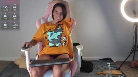 Media: Video of a middle-aged woman with short brown hair, wearing an oversized orange graphic tee, sitting in a pink gaming chair. She holds a gaming controller, surrounded by a cluttered room with a ring light and a stuffed animal.