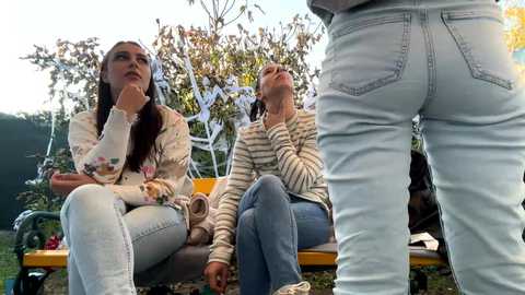 Media: Video of three women in casual attire, sitting on a bench in a garden; one woman stands behind them, wearing tight jeans, creating a playful dynamic.