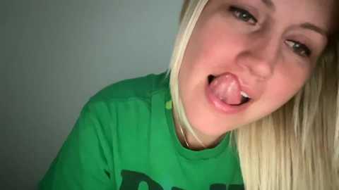 Media: Video of a young, fair-skinned, blonde woman with straight hair, wearing a green t-shirt, sticking her tongue out playfully against a plain background.