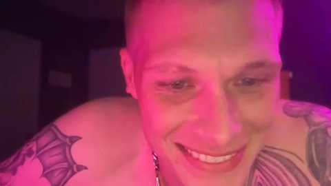 Media: Video of a bald, smiling man with tattoos on his shoulders, bathed in pink and purple lighting.