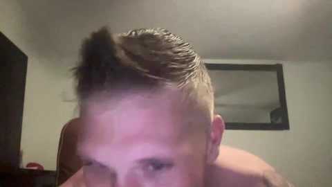 Media: Video of a young man with short, slicked-back hair and a large, noticeable mohawk, taken from a low angle. He has a light skin tone and is shirtless. Background shows a plain room with a mirror and part of a door.