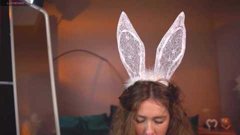 Media: Video of a young woman with long, wavy brown hair, wearing white lace bunny ears, in a dimly lit room with a large, round, orange light fixture in the background.