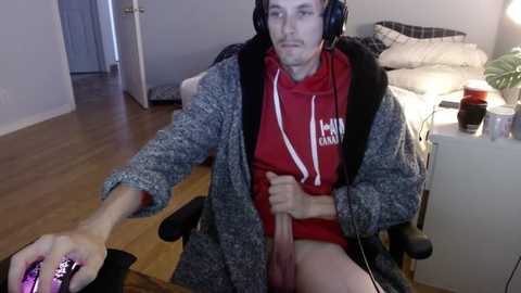 Media: Video of a young man in a gaming chair, wearing headphones and a gray cardigan over a red hoodie, holding a controller, with a visible erect penis. Background shows a cluttered bedroom with a bed, a lamp, and a partially open door.