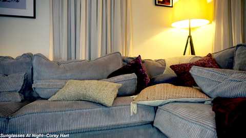 Media: A cozy living room with a light gray sectional sofa adorned with various patterned and textured throw pillows. A tall, floor lamp with a yellow lampshade provides warm lighting against cream-colored curtains and a white wall.