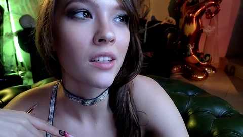 Media: Video of a young woman with light skin, brown hair, and blue eyes, wearing a sparkling choker necklace and a green bra, seated on a dark leather couch. The background includes a green light, a dragon statue, and a colorful lamp.