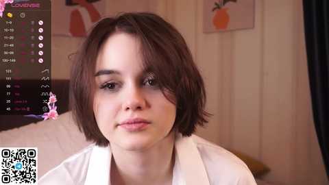 Media: Video of a young woman with shoulder-length brown hair, fair skin, and neutral expression, wearing a white top, in a cozy room with orange wall art and a bed in the background.