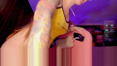 Media: Video of a woman with a tattooed arm holding a pink phone, standing in a dimly lit room with purple lighting.