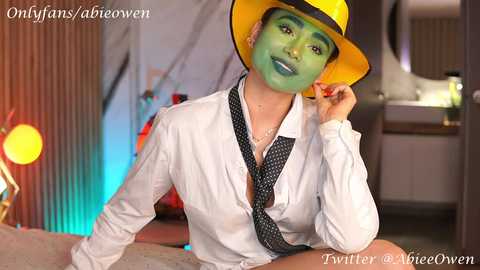 Media: Video of a woman in a white button-up shirt and polka-dot tie, wearing a green face mask, a yellow fedora, and blue lipstick, seated indoors with colorful ambient lighting, watermarked with social media handles.