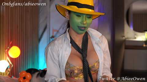 Media: Video of a woman in a yellow fedora, green face paint, and a floral bra, standing in front of a retro-style backdrop with colorful lights.