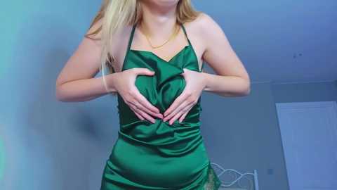 Media: Video of a fair-skinned, blonde woman with a slim figure, wearing a green satin halter dress, hands on her abdomen, in a dimly lit bedroom with white walls and a bed visible in the background.