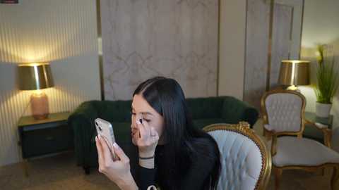 Media: A video of a woman with long black hair, wearing a black top, playing with a white phone. She sits on a green couch, surrounded by a beige wall with floral patterns, a gold lamp, and a white chair with a gold frame.