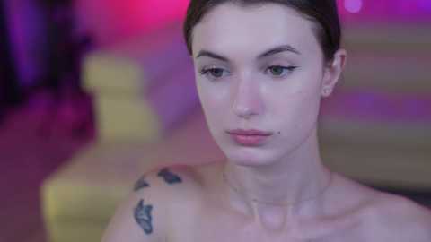 Media: Video of a fair-skinned woman with brown hair, wearing minimal makeup, and a small tattoo on her right shoulder. She has a contemplative expression, set against a softly blurred background with pink and purple hues.