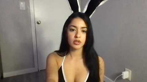 Media: Video of a Latina woman with long black hair, wearing a revealing black bunny costume with white trim, posing indoors with gray walls and a white door.