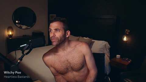 Media: Video of shirtless, muscular, bearded man with short hair, seated on a bed in a dimly lit bedroom, looking thoughtful.