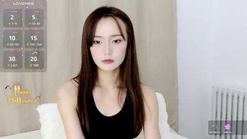 Media: Video of an East Asian woman with long, straight brown hair, wearing a black tank top, sitting on a white bed. Background features a calendar and a radiator.