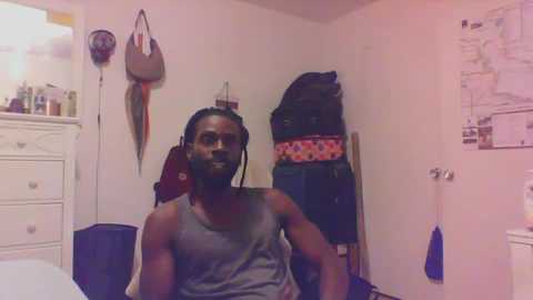 Media: A video of a muscular, dark-skinned man with dreadlocks in a gray tank top, sitting in a cluttered bedroom with a white dresser, hanging bags, and a colorful wall poster.