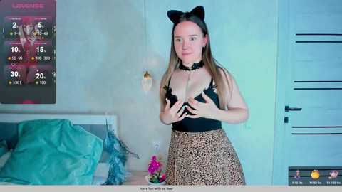 Media: Video of a light-skinned woman with long brown hair, wearing black lingerie with leopard-print skirt, cat ears, and black choker, standing in a modern bedroom with blue walls and a calendar, holding her breasts.