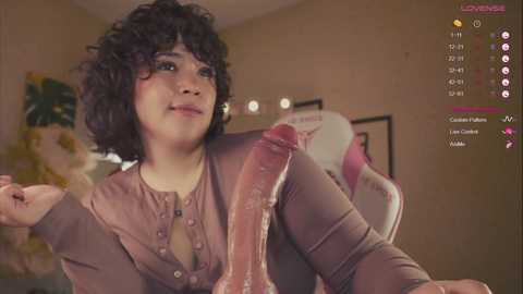 Media: A digital image featuring a curvy, light-skinned woman with curly black hair and medium breasts, wearing a pink blouse. She holds a large, erect penis, set against a beige wall with a yellow plant in the background.