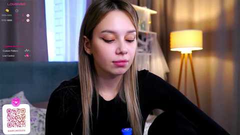 Media: Video of a young woman with long blonde hair, closed eyes, wearing a black top, sitting on a couch in a cozy room with a lamp and window.