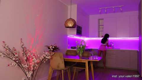 Media: Video of a modern kitchen with a woman in pink, standing at the counter, under purple LED lighting. Wooden table with chairs, brown vase with flowers, and hanging pendant lamp.