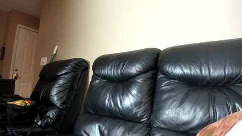 Media: A video of a living room featuring two black leather reclining chairs and beige walls. The chairs are positioned side by side, facing a closed white door. A brown couch is partially visible in the foreground.