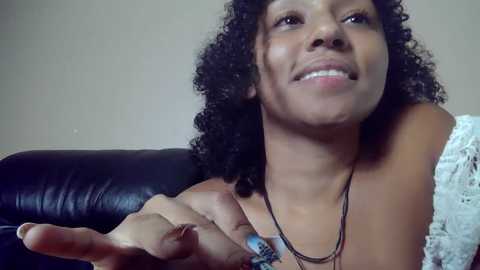 Media: Video of a smiling, light-skinned African-American woman with curly hair, wearing a white lace top and multiple layered necklaces, extending her hand towards the camera. She sits on a black leather couch against a plain beige wall.