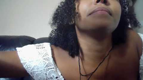 Media: Video of a Black woman with medium-dark skin, curly black hair, and a white lace top, leaning back on a black leather couch.