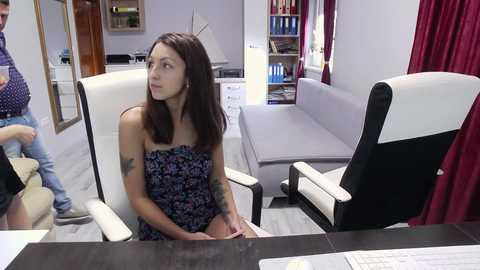 Media: Video of a slender, fair-skinned brunette woman in a strapless floral dress, sitting in a modern office chair, looking contemplative. Background includes a gray couch, bookshelves, and a wooden door.