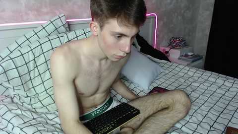 Media: Video of a shirtless, lean, young Caucasian man with short brown hair, wearing green Calvin Klein boxers, typing on a keyboard, in a bed with white checkered sheets, in a dimly lit bedroom.