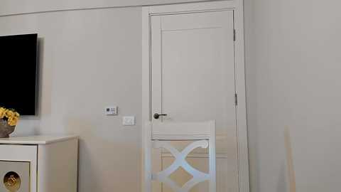 Media: A minimalist room with a white door, beige walls, a white chair, and a flat-screen TV on the left, a flower vase on the white dresser, and a light switch on the wall.