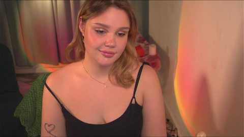 Media: Video of a young woman with light skin, light brown hair, wearing a black spaghetti-strap top, smiling, in a colorful, softly lit room with a heart tattoo on her upper arm.