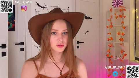 Media: Video of a young, fair-skinned woman with long brown hair, wearing a brown cowboy hat and sleeveless brown top, standing indoors with Halloween decorations.