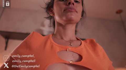 Media: Video of a woman with fair skin and dark hair, wearing an orange top with a large cutout revealing her cleavage. She has a gold necklace and a neutral expression.