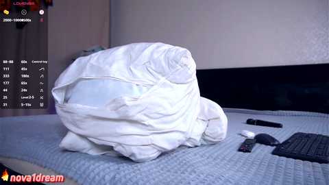 Media: A video showing a white comforter wrapped around a person's head on a bed, with a TV, remote, and keyboard nearby. The room's background features a plain wall and a wooden door.