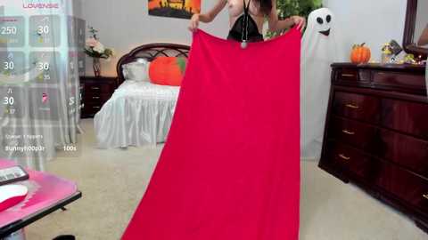 Media: Video of a woman in a black top holding a large, vibrant red blanket in a cozy bedroom with a bed, wooden dresser, and pumpkin decorations.