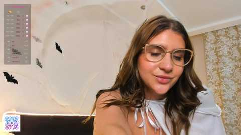 Media: Video of a young woman with light skin and brown hair wearing large, round glasses, a white blouse, and a subtle smile. Background features a white ceiling with black bat decals and a floral-patterned curtain.