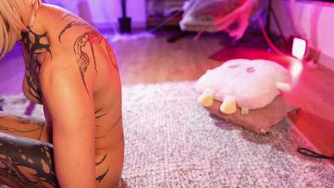 Media: Video of a topless, tattooed, blonde woman kneeling on a plush carpet in a dimly lit room, with a large, pink plush toy and a red lamp in the background.