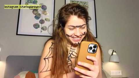 Media: Video of a smiling, topless woman with long brown hair, wearing black spider web makeup, holding an iPhone, in a bedroom with framed artwork on the wall.