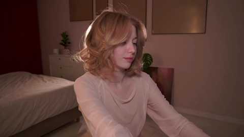 Media: A video of a young woman with light skin and shoulder-length, wavy blonde hair, wearing a cream-colored top, sitting on a bed in a dimly lit, minimalist bedroom.