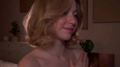 Media: Video of a smiling, light-skinned woman with short, wavy blonde hair, wearing a delicate necklace. She holds a green leaf, standing in a dimly lit bedroom with a white dresser and potted plant in the background.